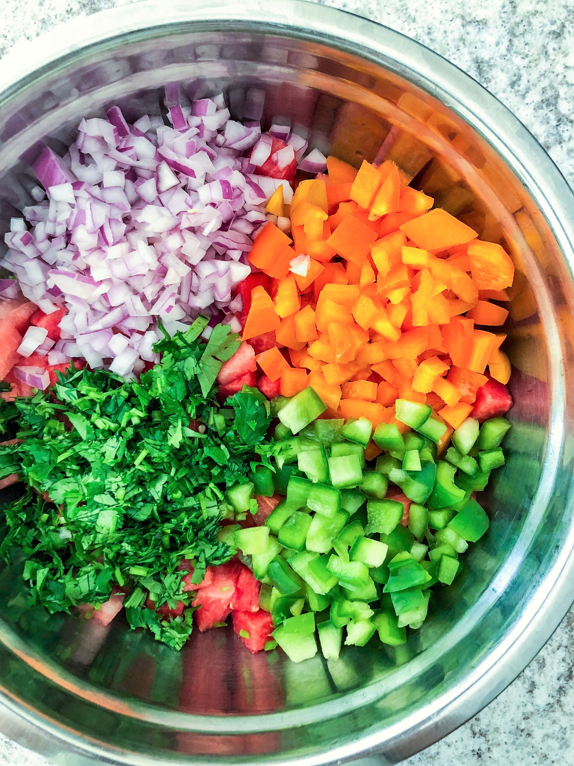 Ingredients of watermelon jalapeno salsa in a stainless steel mixing bowl include orange and green bell peppers, red onions, jalapenos, and cilantro.