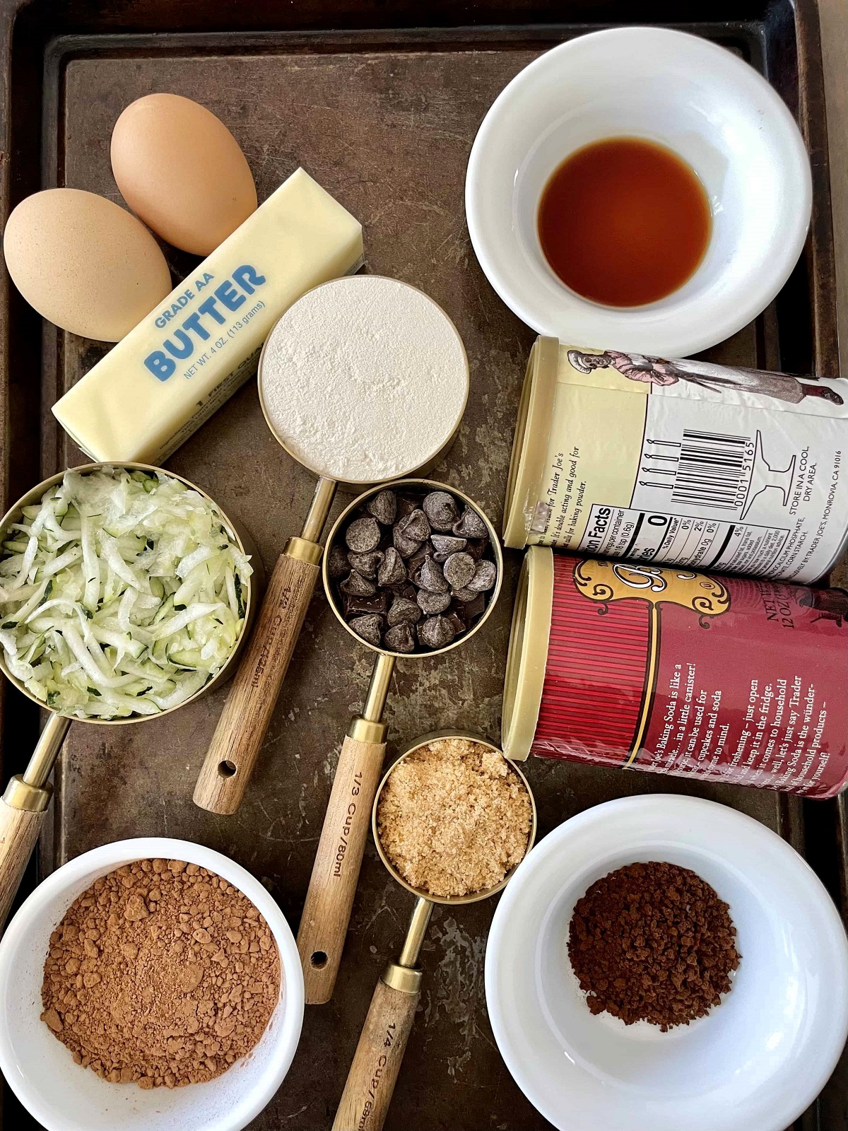 butter, eggs, flour, zucchini, vanilla, baking powder and soda, coffee granules, cocoa powder, and brown sugar on a metal tray.