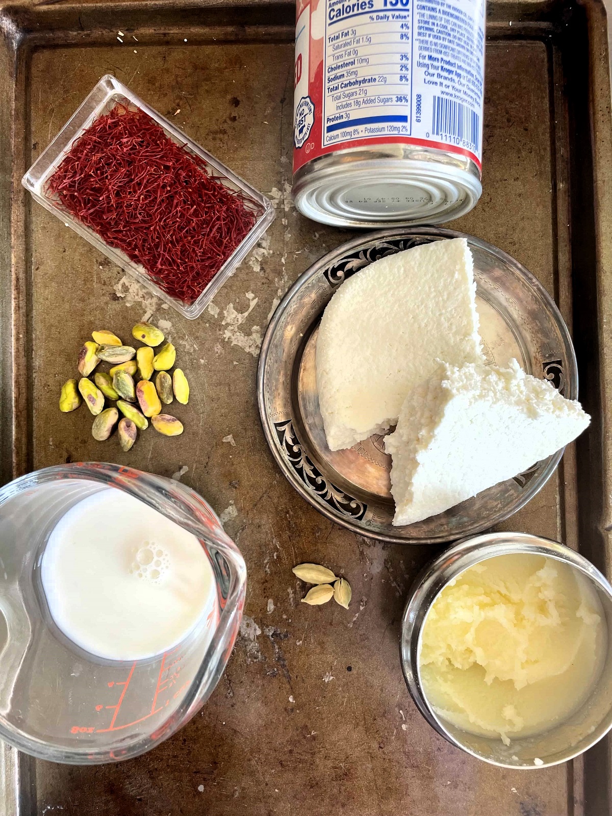 Saffron, pistachios, milk, cardamom and other Peda recipe ingredients on a metal cookie sheet.