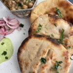 Kulcha Bread sprinkled with chopped cilantro served with green chtney, pickled red onions, and chickpea curry in a bowl on a ceramic plate.