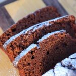 Close up of baked coffee and chocolate loaf cake dusted with powdered sugar on a wooden board.