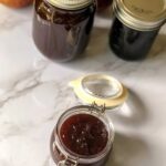 Apple butter in a small jar with an open lid and more jars of apple butter in the background on a white marble countertop.