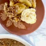 Apple crisp served on a terracotta plate along with a scoop of vanilla ice cream.