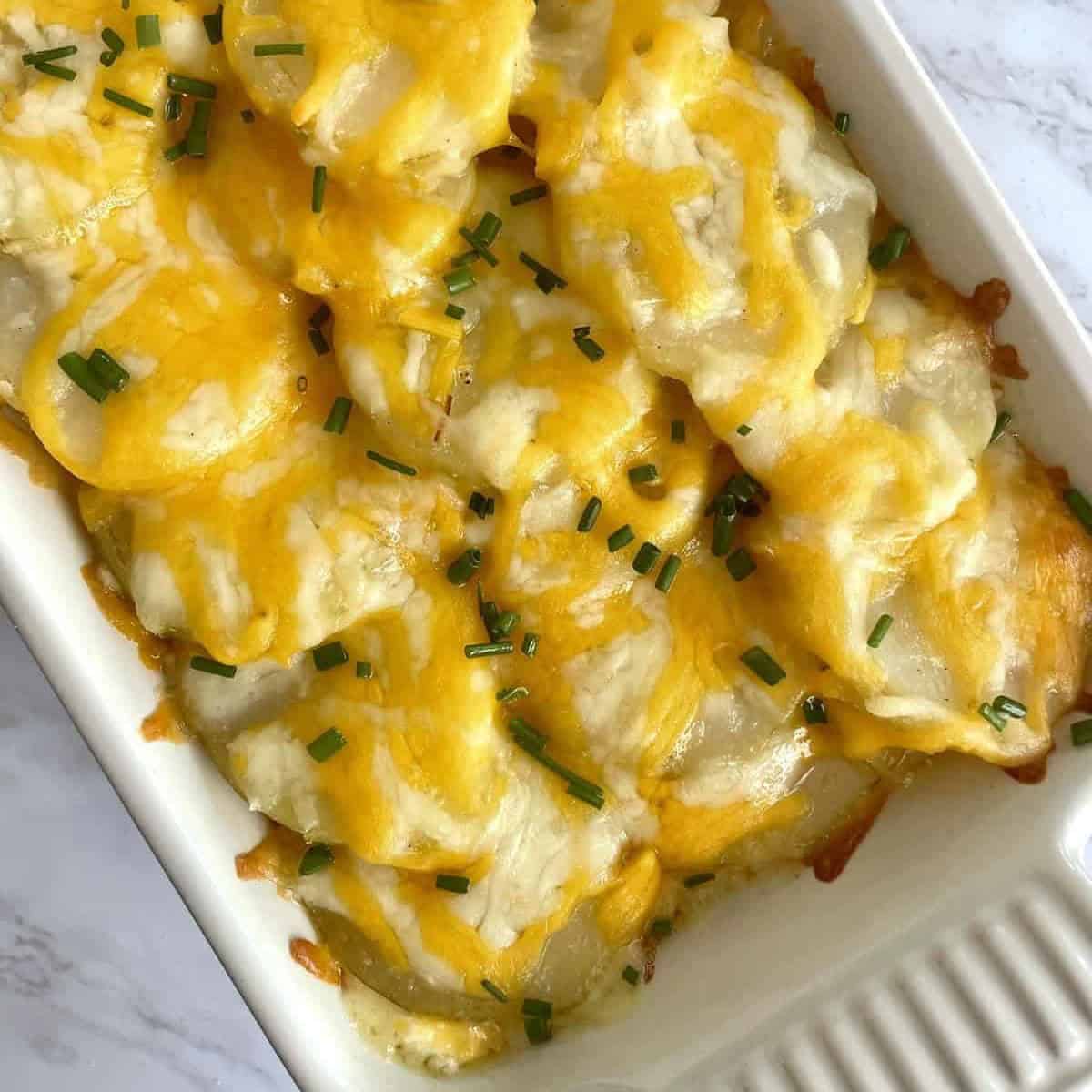 Baked Potato Casserole with cheese and chopped chives in a white ceramic dish on a white marble background.