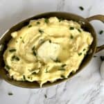 Mashed Potatoes with a pat of butter and garnished with chopped chives in a steel serving dish on a white marble background.
