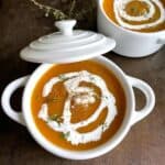 Butternut squash soup in a white ceramic bowl on a dark metal cookie sheet. The soup is drizzled with cream and garnished with thyme leaves and fresh black pepper.