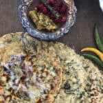 Three methi wali makki ki rotis with melting butter on a dark cookie sheet with chili peppers and a bowl of green chili and cranberry pickle.