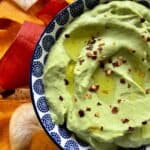 Avocado cottage cheese dip in a blue and white ceramic bowl with red pepper flakes and olive oil set in a ceramic plate full of colorful chips.