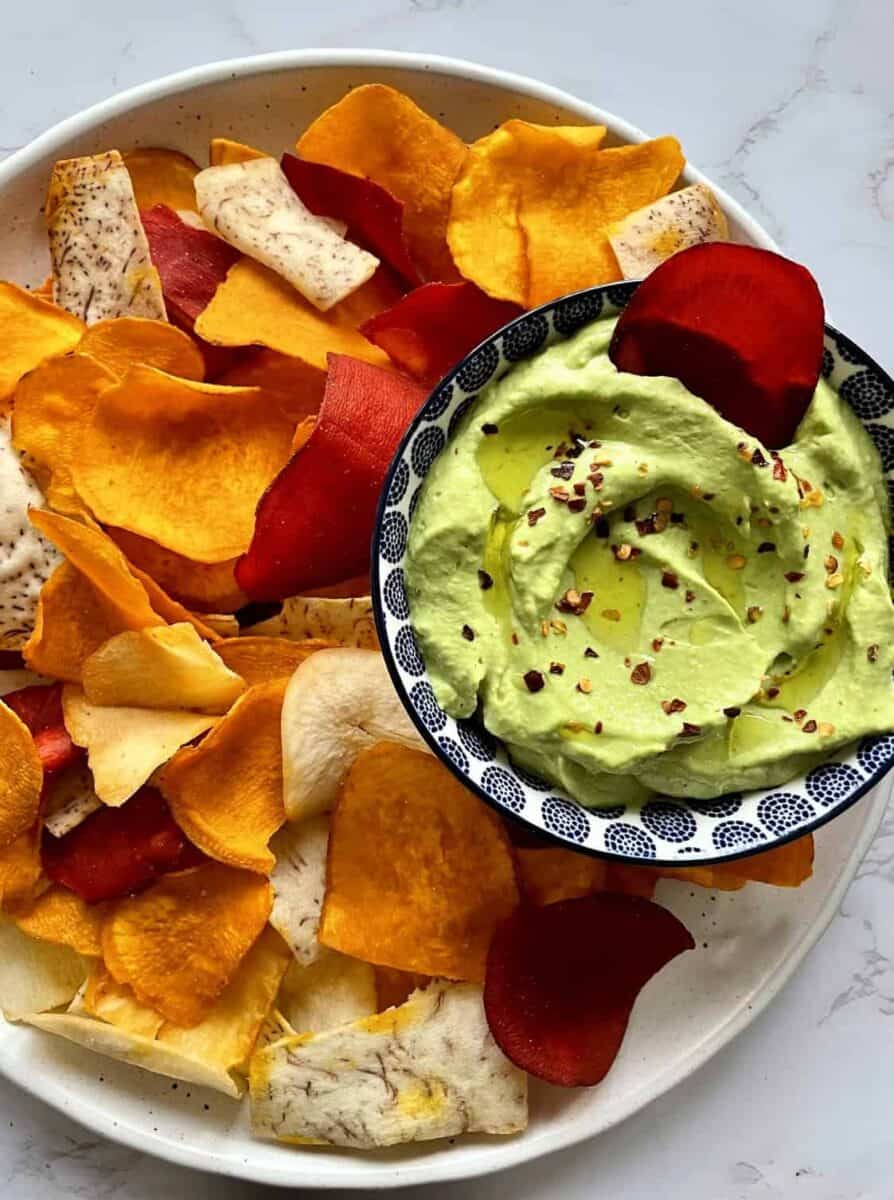 Avocado cottage cheese dip in a blue and white ceramic bowl with red pepper flakes and olive oil set in a ceramic plate full of colorful chips.