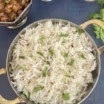 Cooked rice with cumin seeds and garnished with chopped cilantro in a stainless steel bowl with gold handles on a blue backdrop. A bowl of chickpea curry and a bowl of red onion salad in another bowl in the background.