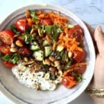 A bowl with cottage cheese, chopped veggies, seasoning, peanuts, and chopped cilantro held in a hand with gold bangles over a white marble background.