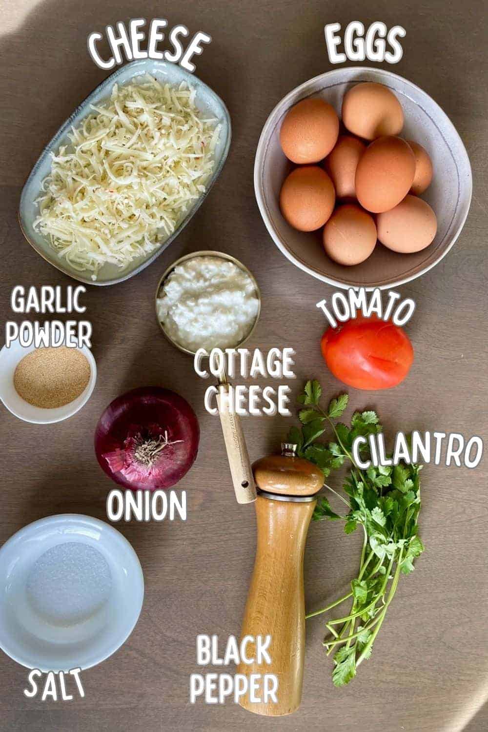 Ingredients for baked egg bites on a wooden background including eggs, cottage cheese, salt, pepper, garlic powder, cilantro, tomato, onion, and cheese.