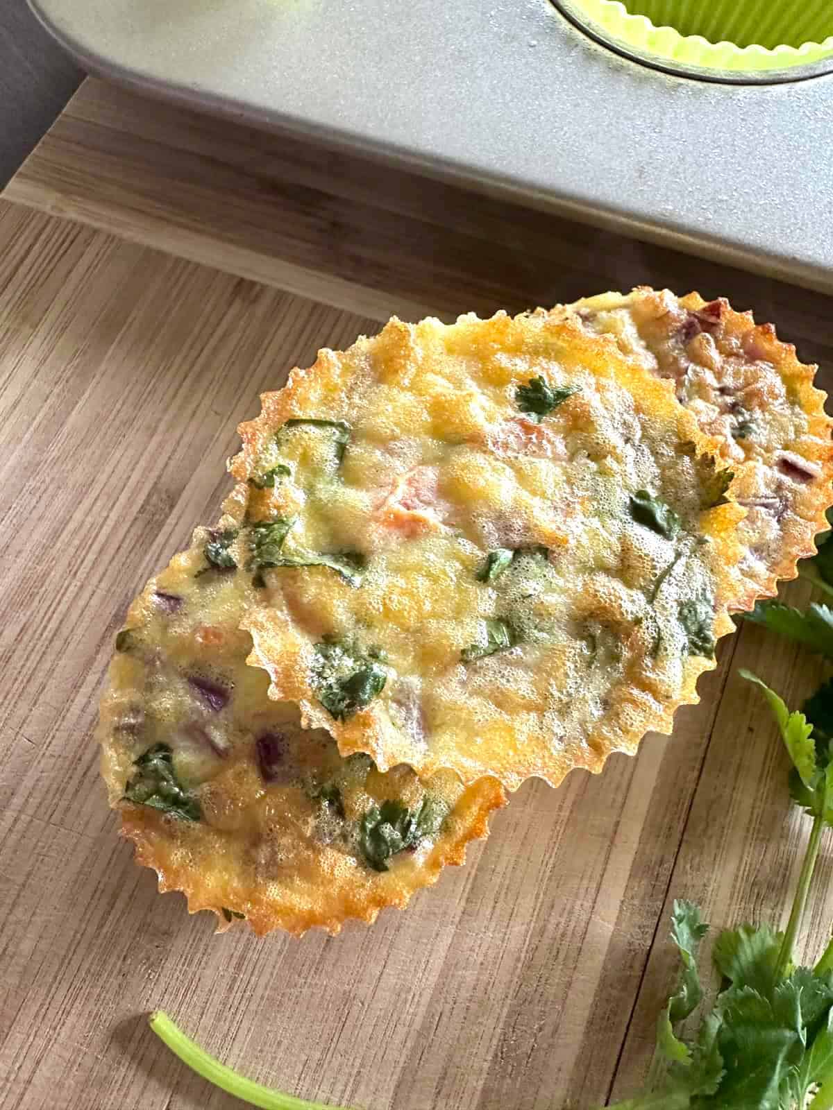 Baked egg bites with onion, cilantro and tomato stacked on top of two other egg bites on a wooden chopping block.