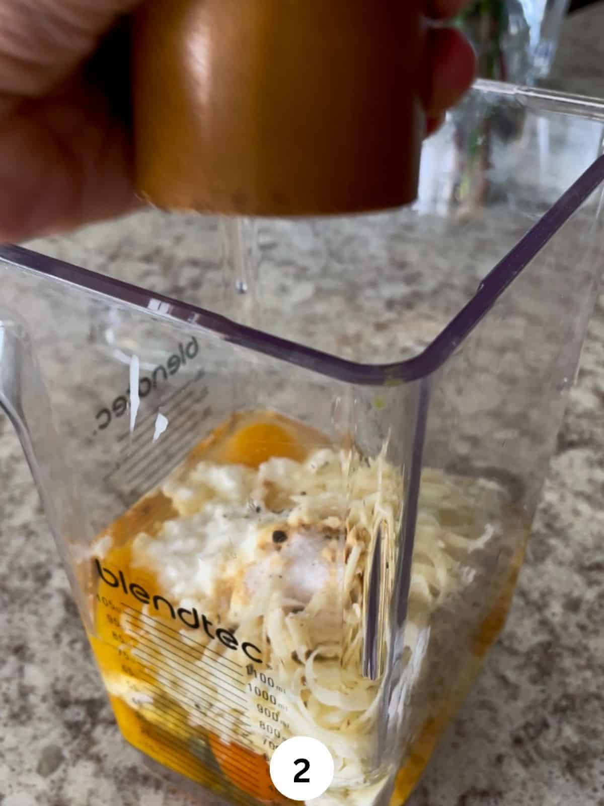 Egg whites and yokes, cottage cheese, and shredded cheese in the jar of a blender along with seasoning and black pepper coming from a handheld pepper mill.