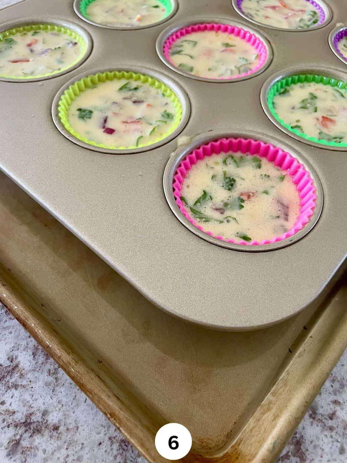 Egg and cheese mixture with chopped cilantro, onions, and tomatoes in a lined muffin pan kept in a cookie sheet with water in it.
