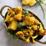 Potato and cauliflower in an oval stainless steel serving dish garnished with cilantro on a white marble background with lemon wedges and cilantro sprigs in the background.