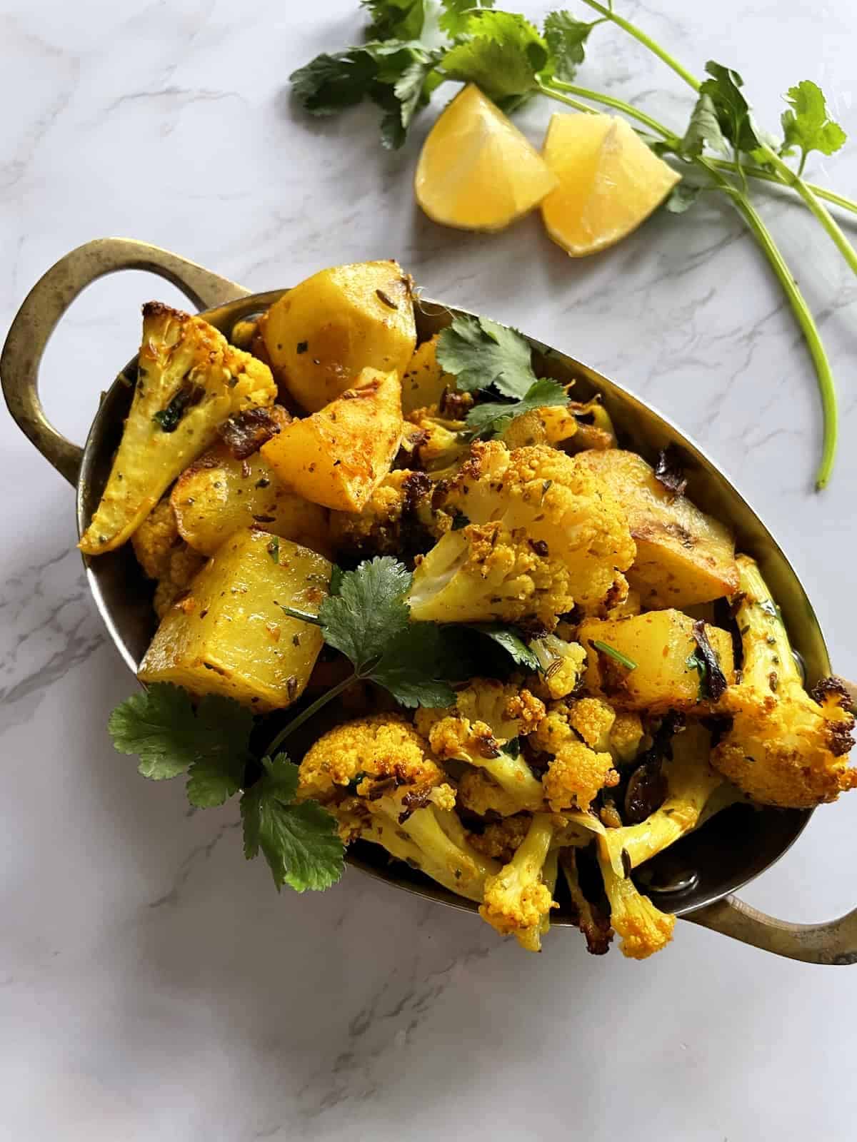Potato and cauliflower in an oval stainless steel serving dish garnished with cilantro on a white marble background with lemon wedges and cilantro sprigs in the background.