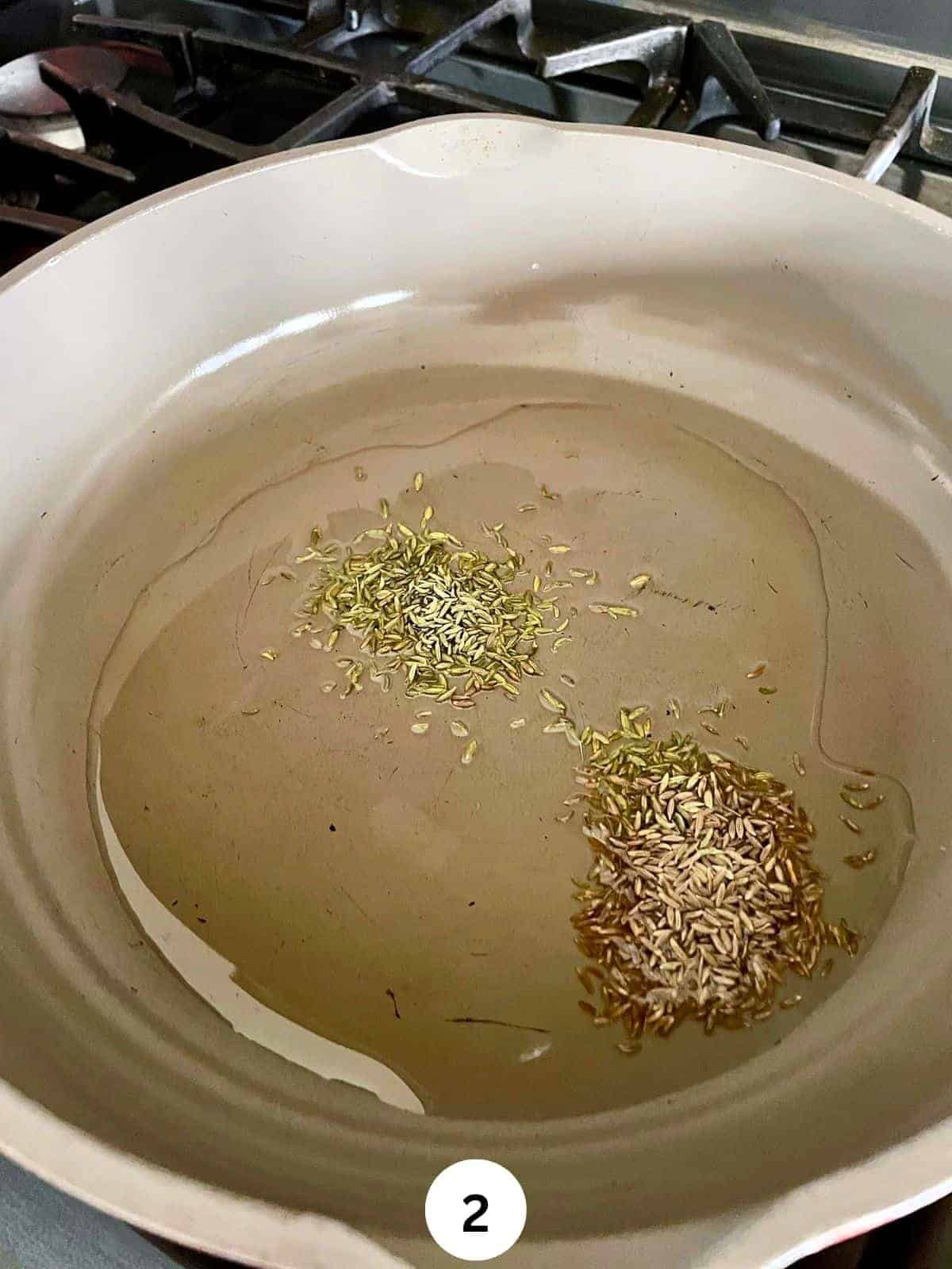 Oil in a grey ceramic saute pan with cumin seeds and fennel seeds.