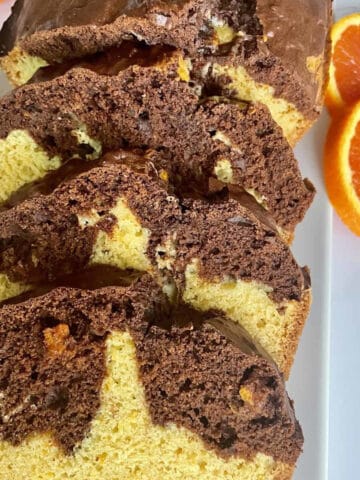 A marbled chocolate and orange loaf cake sliced and arranged on a white rectangular ceramic platter with slices of orange along the sides of the platter for garnish.