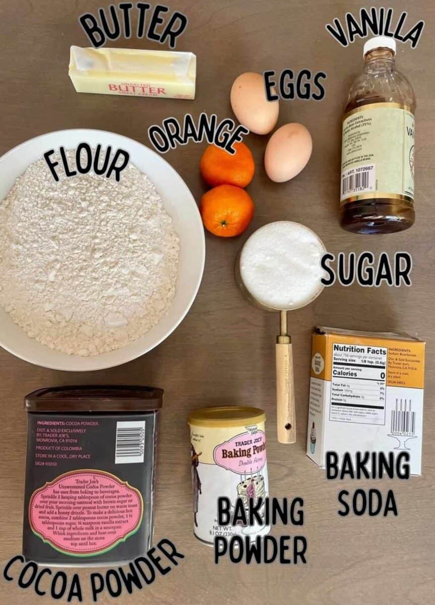 Ingredients for making chocolate orange loaf cake on a wooden background including flour, sugar, butter, eggs, oranges, cocoa powder, and vanilla.