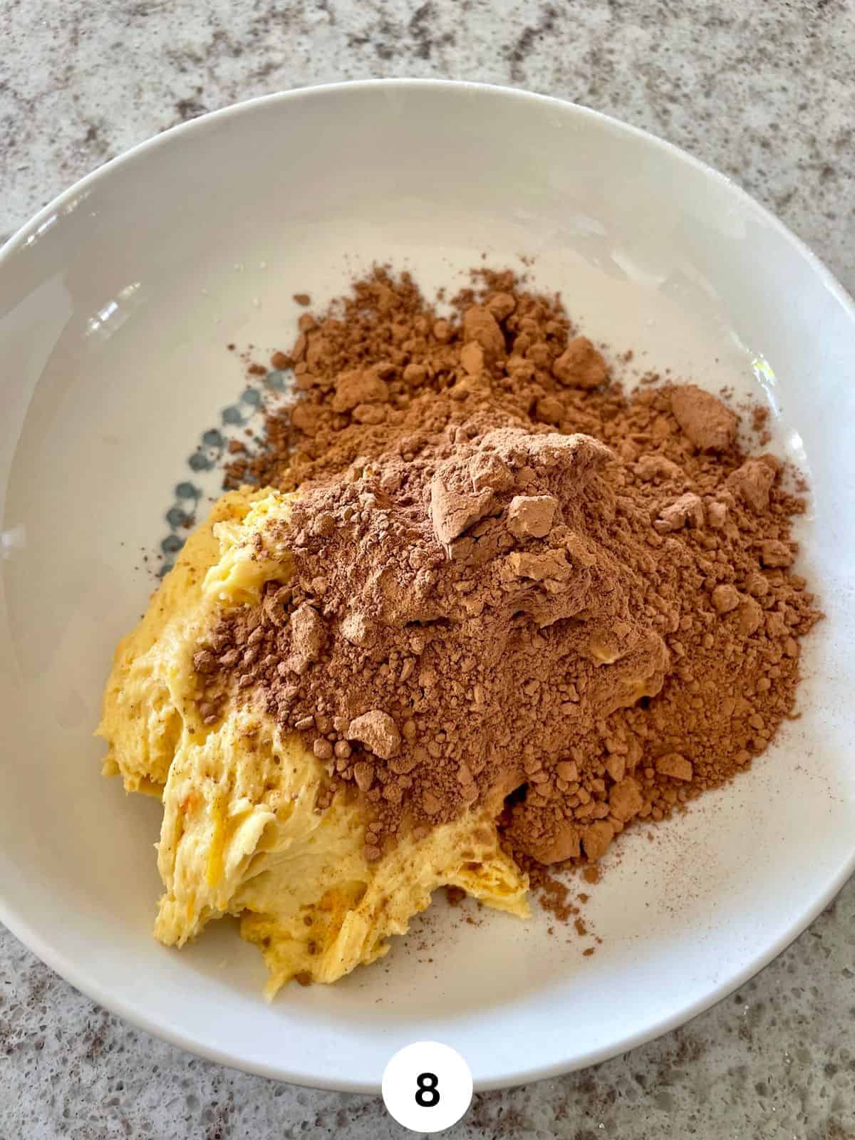 Orange cake batter with cocoa powder sprinkled over it in a wide round ceramic bowl.