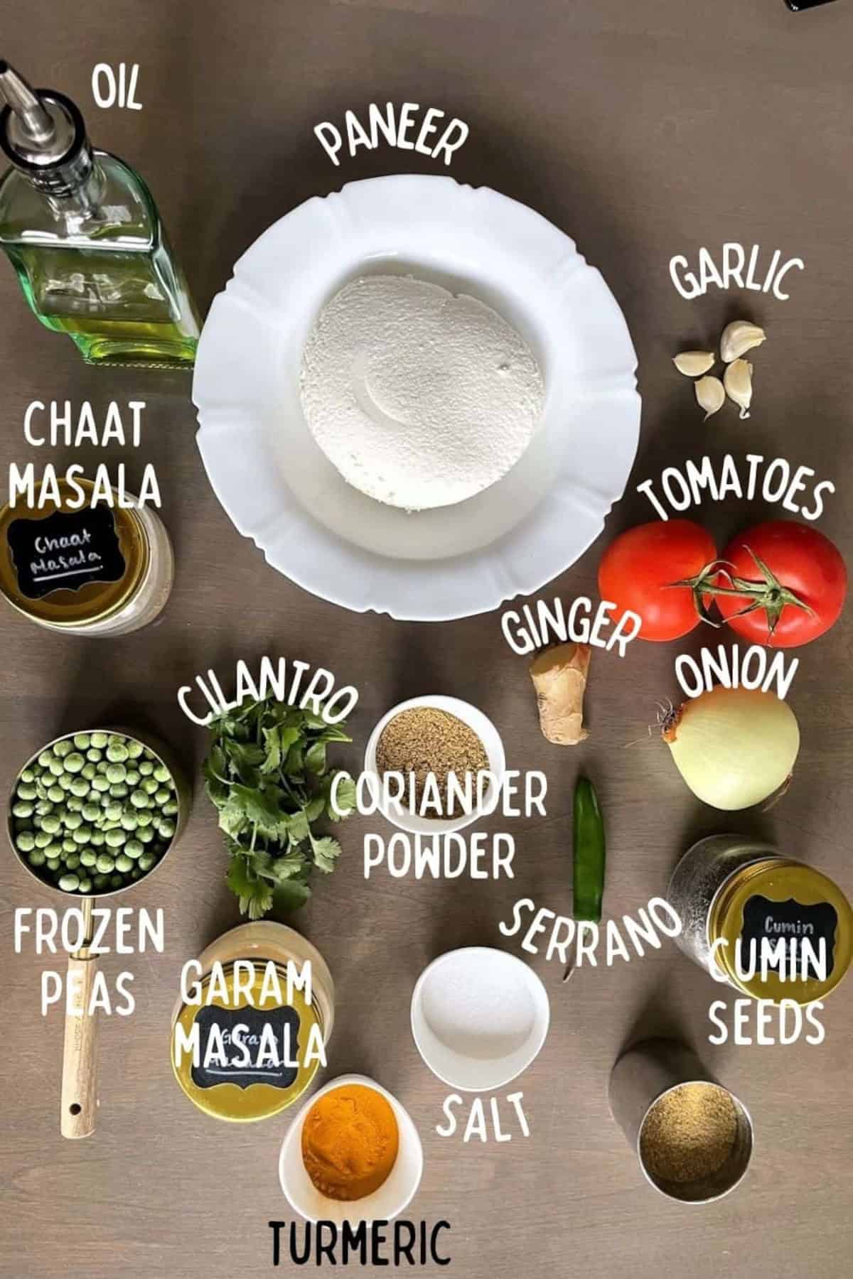 Ingredients of Paneer bhurji on a wooden background. The ingredients include paneer, onion, tomatoes, ginger, garlic, frozen peas, oil, and spices.