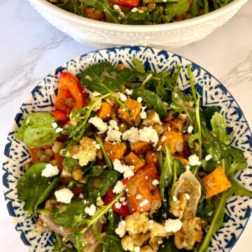 A lentil and roasted vegetable salad with spinach and feta cheese served in a white and blue ceramic bowl against a white marbled background.
