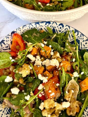 A lentil and roasted vegetable salad with spinach and feta cheese served in a white and blue ceramic bowl against a white marbled background.
