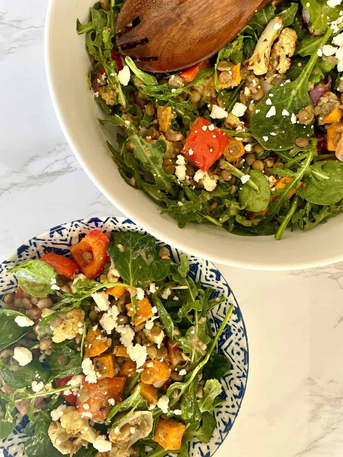 A lentil and roasted vegetable salad with spinach and feta cheese served in a white and blue ceramic bowl against a white marbled background.