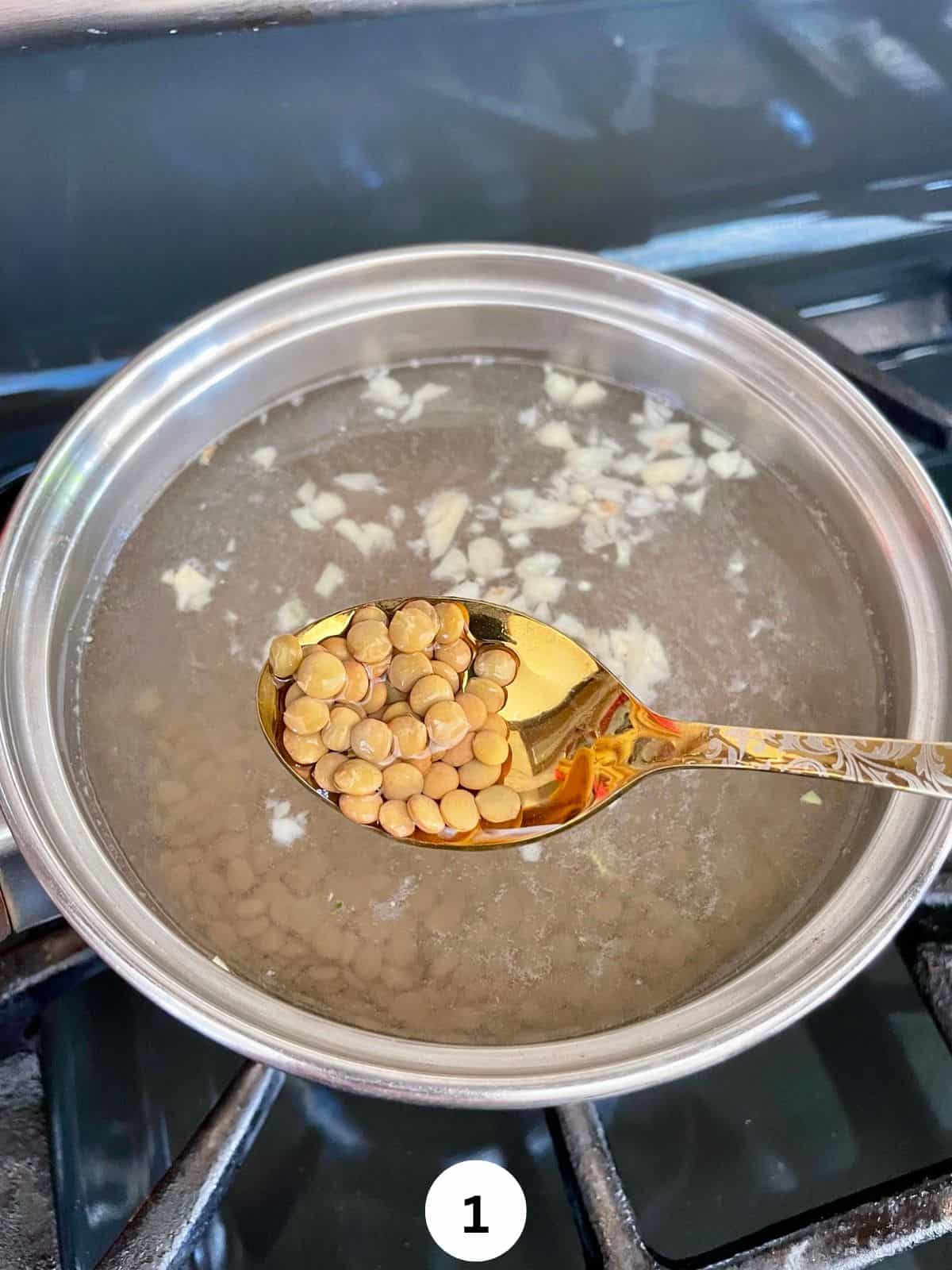 Brown lentils in a spoon held above a sauce pan with the lentils cooking in it.