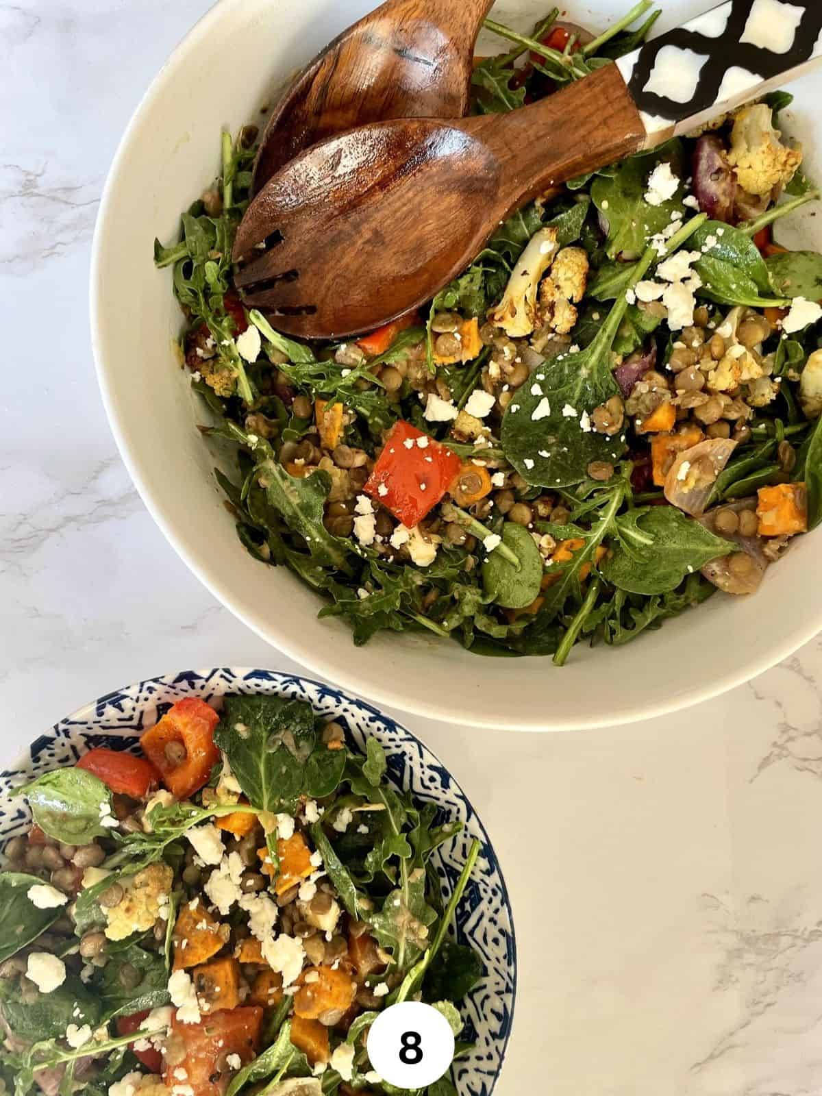A lentil and roasted vegetable salad with spinach and feta cheese served in a white and blue ceramic bowl and a big white serving bowl against a white marbled background.