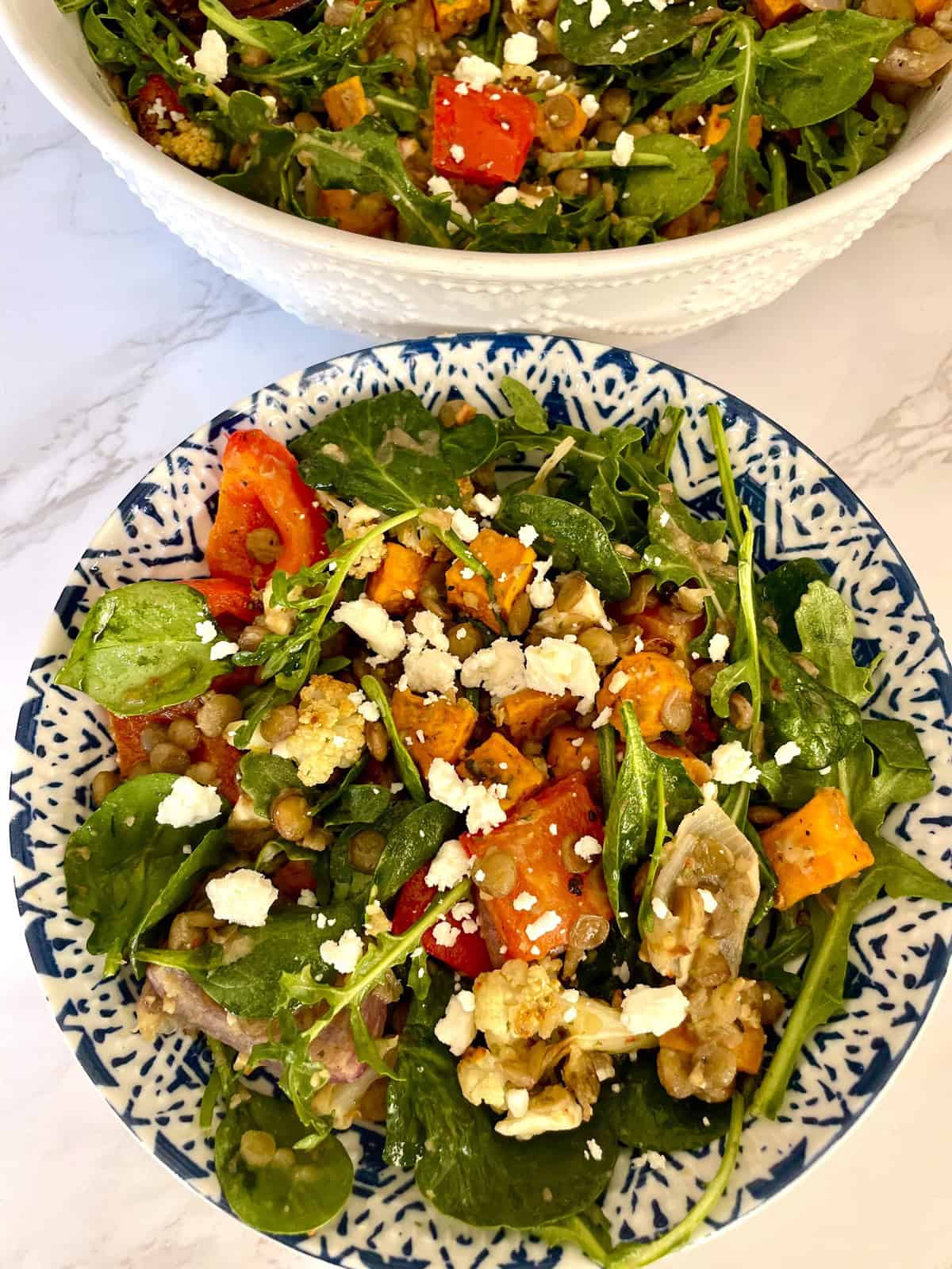 A lentil and roasted vegetable salad with spinach and feta cheese served in a white and blue ceramic bowl against a white marbled background.