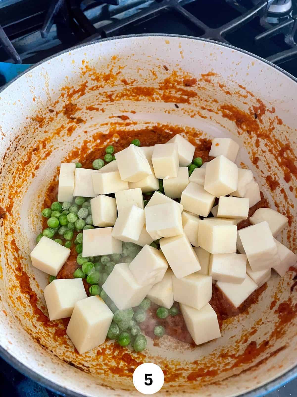 Paneer cubes and frozen peas with cooked masala in the bottom in an enameled cast iron pan.