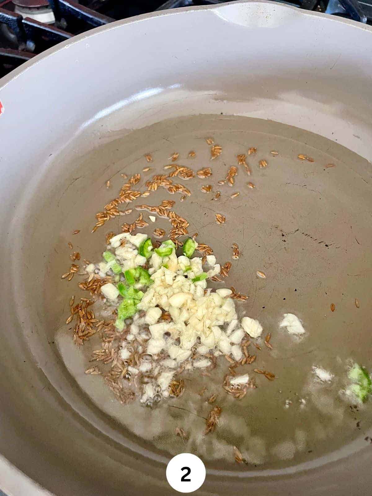 Chopped garlic and green chili sauteing with oil and cumin seeds in a grey ceramic pan.