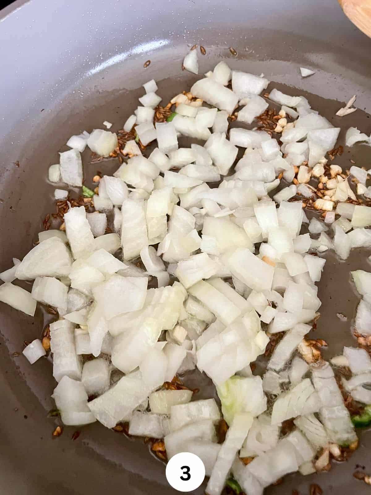 Chopped onions in a grey ceramic pan cooking with oil and cumin seeds.