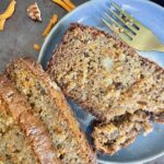 Slice of carrot cake in a blue ceramic plate with a golden fork with some shredded carrots and pecan pieces in the background.
