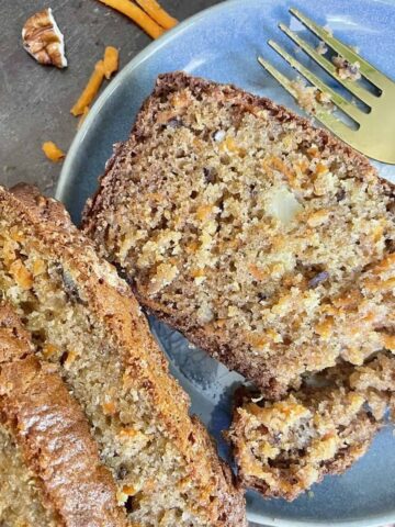 Slice of carrot cake in a blue ceramic plate with a golden fork with some shredded carrots and pecan pieces in the background.