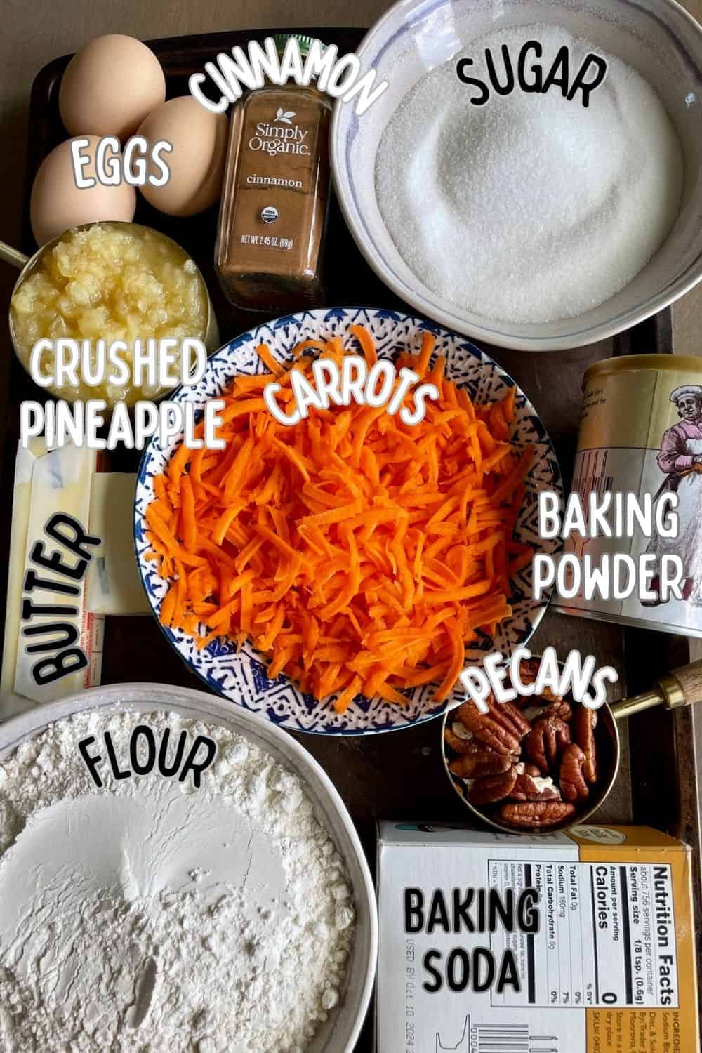 Ingredients of southern carrot cake with pineapple on a dark background, including shredded carrots, crushed pineapple, flour, sugar, baking powder, cinnamon, and baking soda.