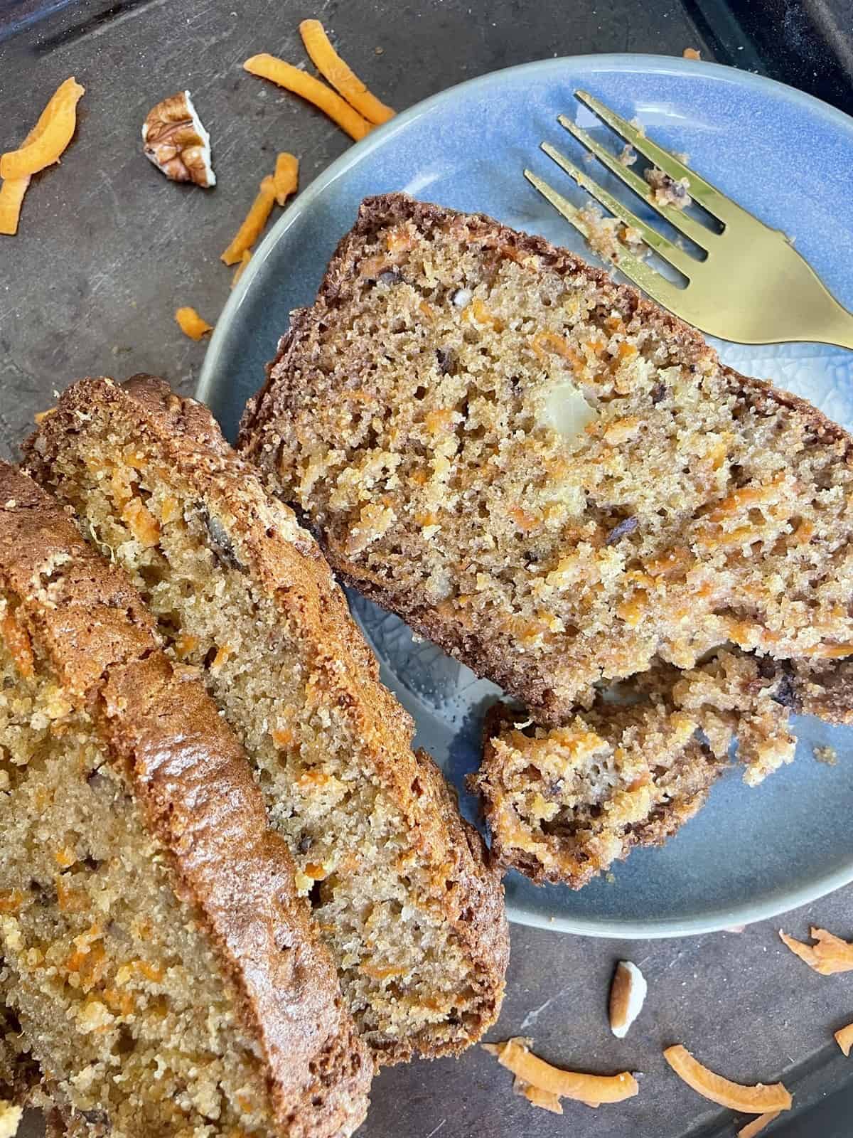 Slice of carrot cake in a blue ceramic plate with a golden fork with some shredded carrots and pecan pieces in the background.