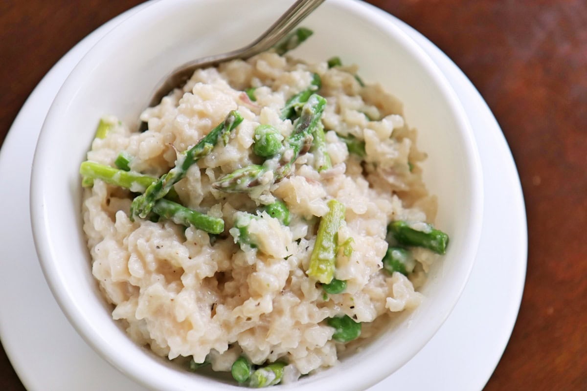 Creamy risotto with asparagus and peas in a white ceramic bowl kept on a plate on a wooden table.
