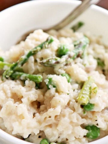 Close up of creamy risotto with asparagus and peas in a white ceramic bowl.