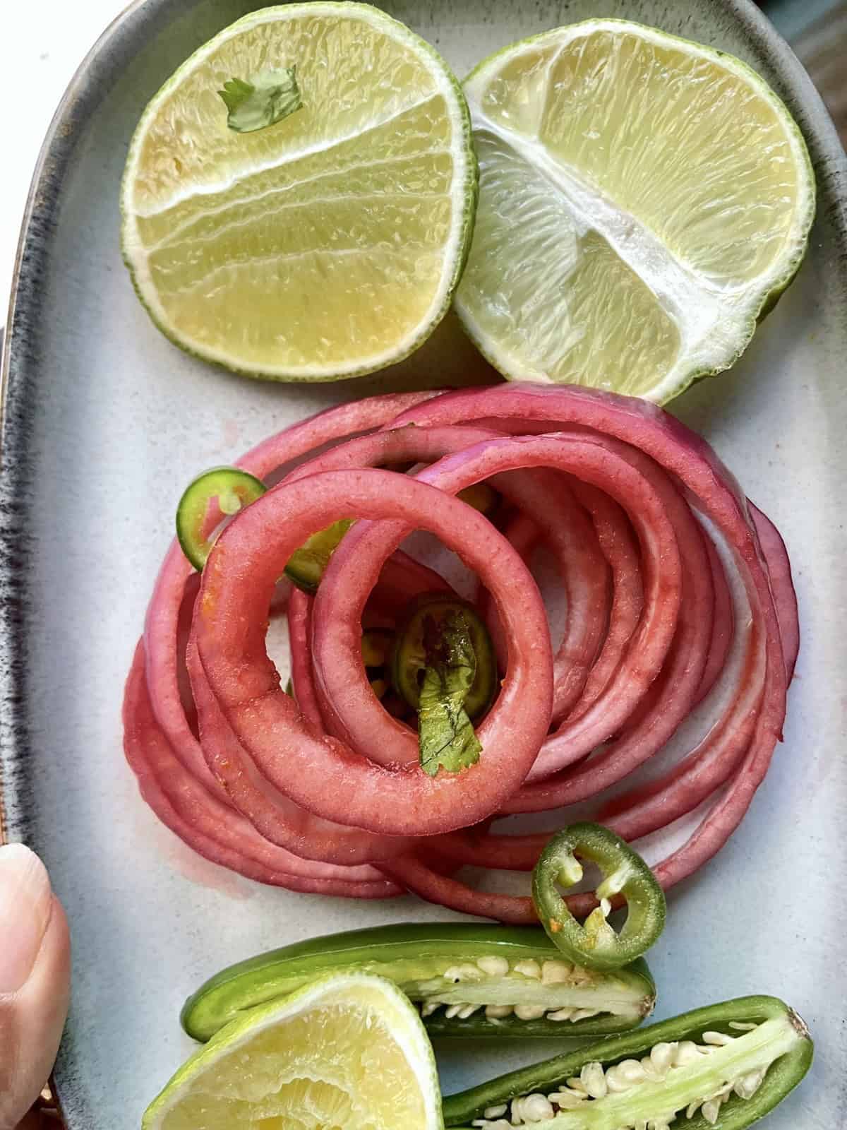 A stack of pickled red onions in a grey ceramic tray with slices limes and serrano peppers.