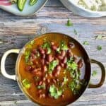 Rajma or red kidney beans curry garnished with chopped cilantro in a stainless steel serving bowl with golden handles on a wooden table. A small ceramic tray with sliced limes, pickled onions, and green chili peppers and rice in a ceramic bowl are right next to the serving dish.