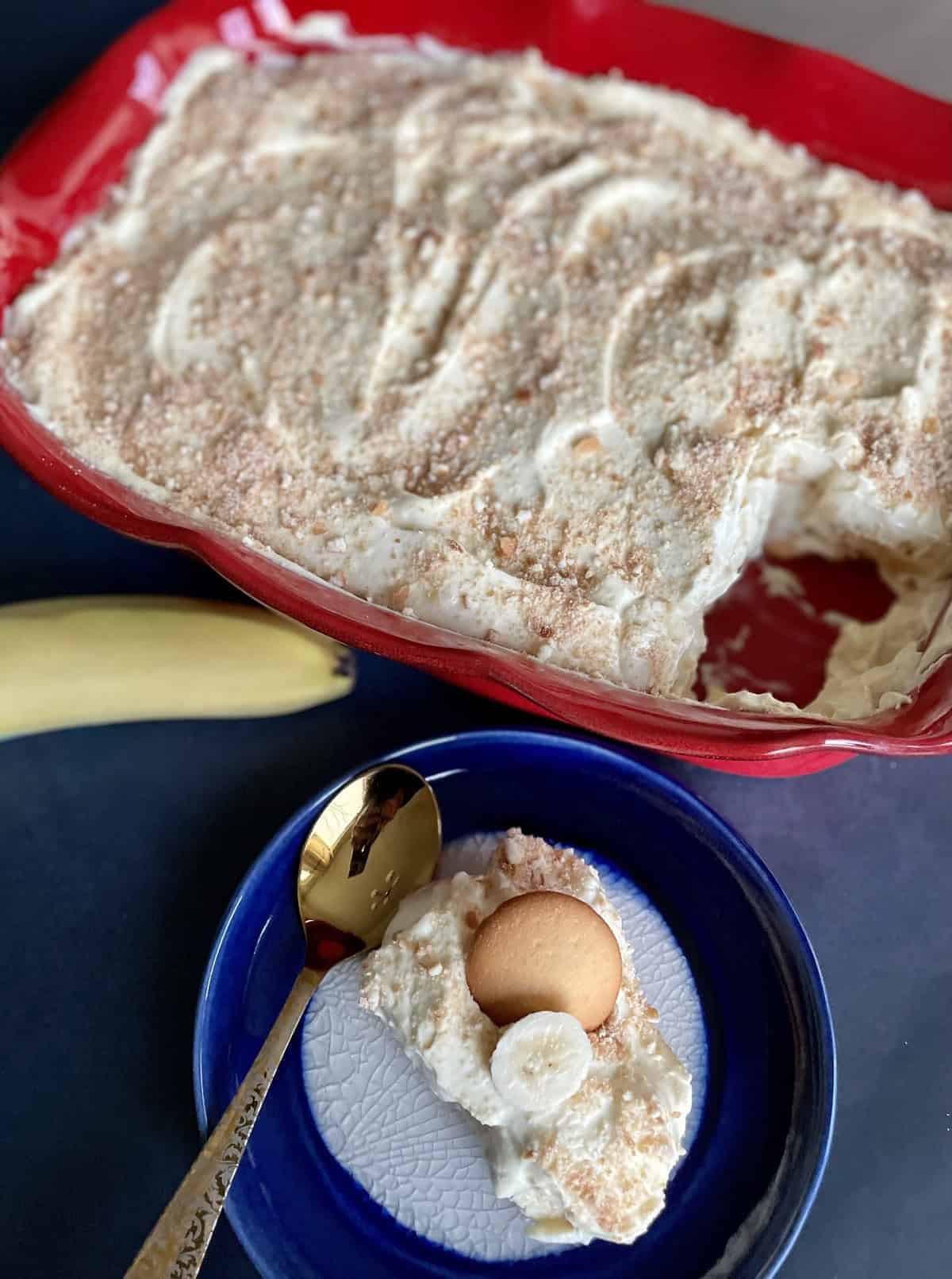 Banana pudding with a vanilla wafer on top in a blue and white ceramic plate on a blue background. A red ceramic dish with fluted edges with banana pudding with a slice taken out.