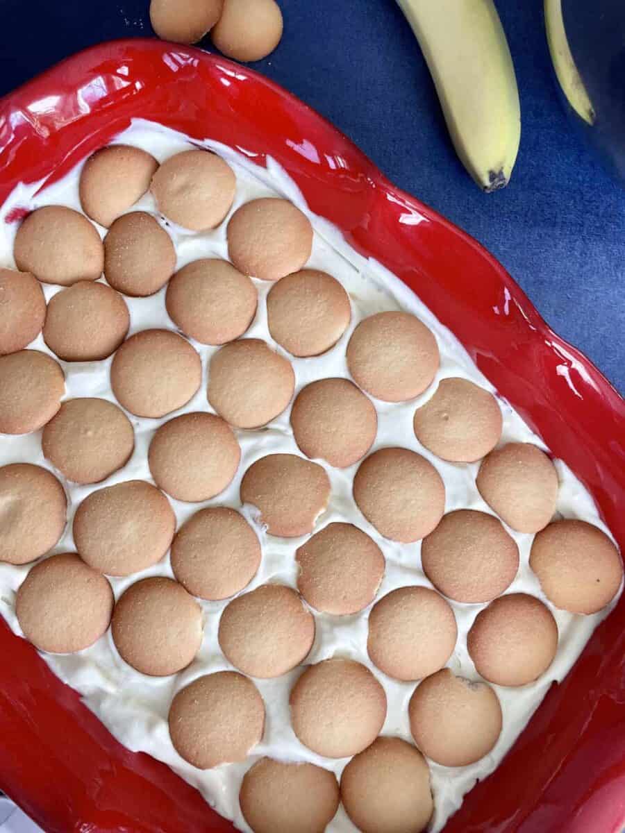 A layer of vanilla wafers sits on top of vanilla pudding layer in a red ceramic dish.