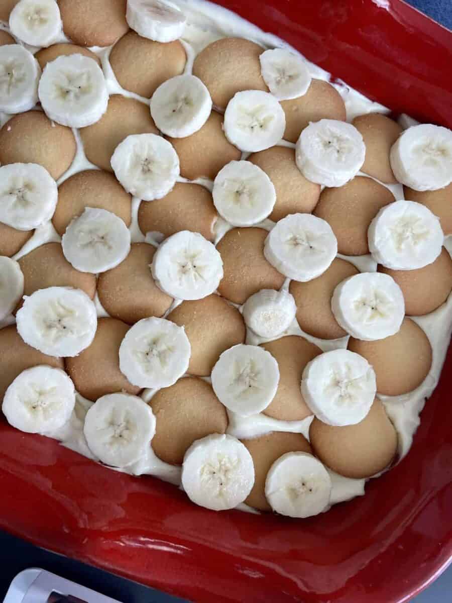 Slices of peeled bananas layered over vanilla wafers and vanilla pudding in a red ceramic dish.