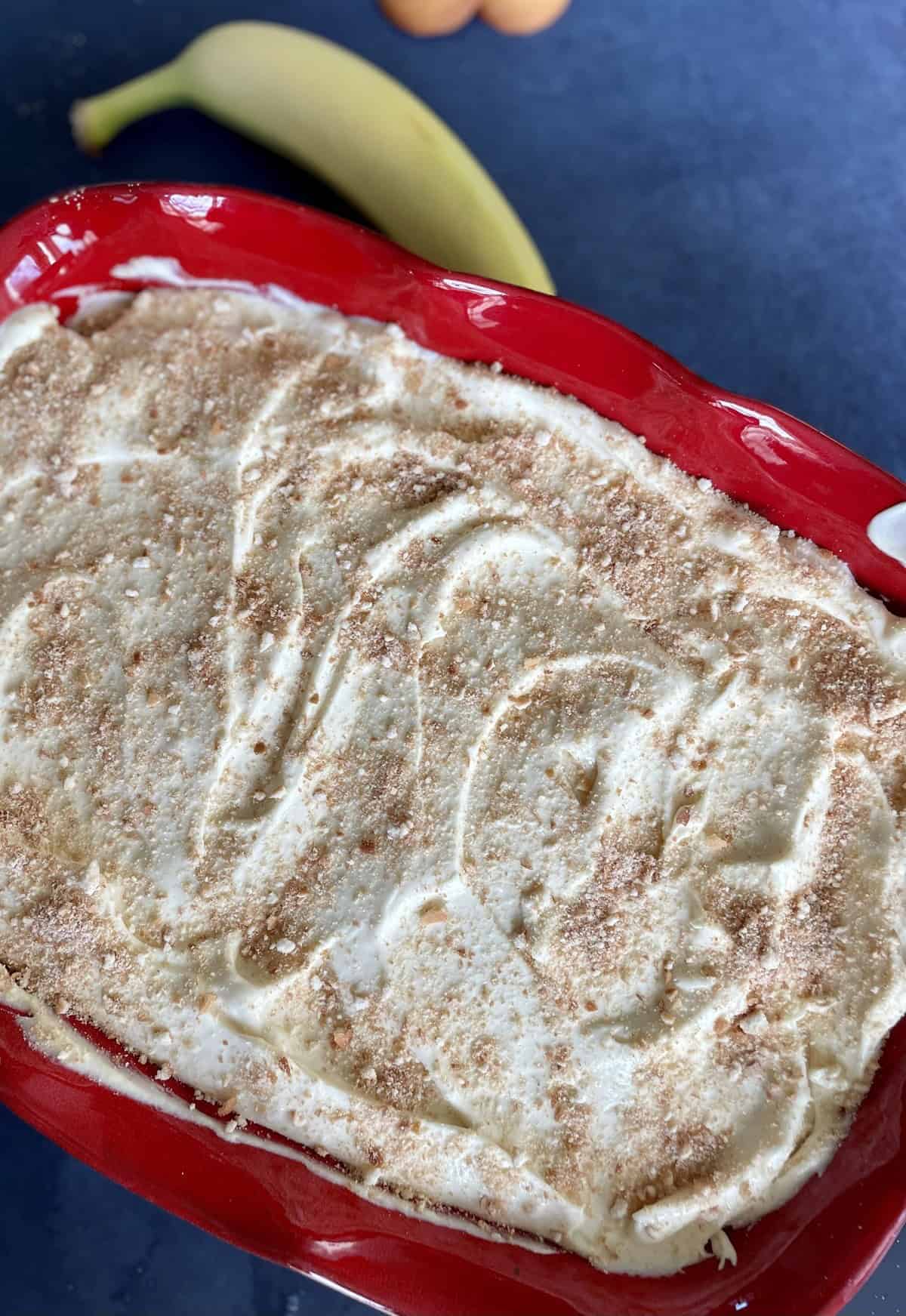 Banana pudding sprinkled with crushed vanilla wafers in a red ceramic dish.