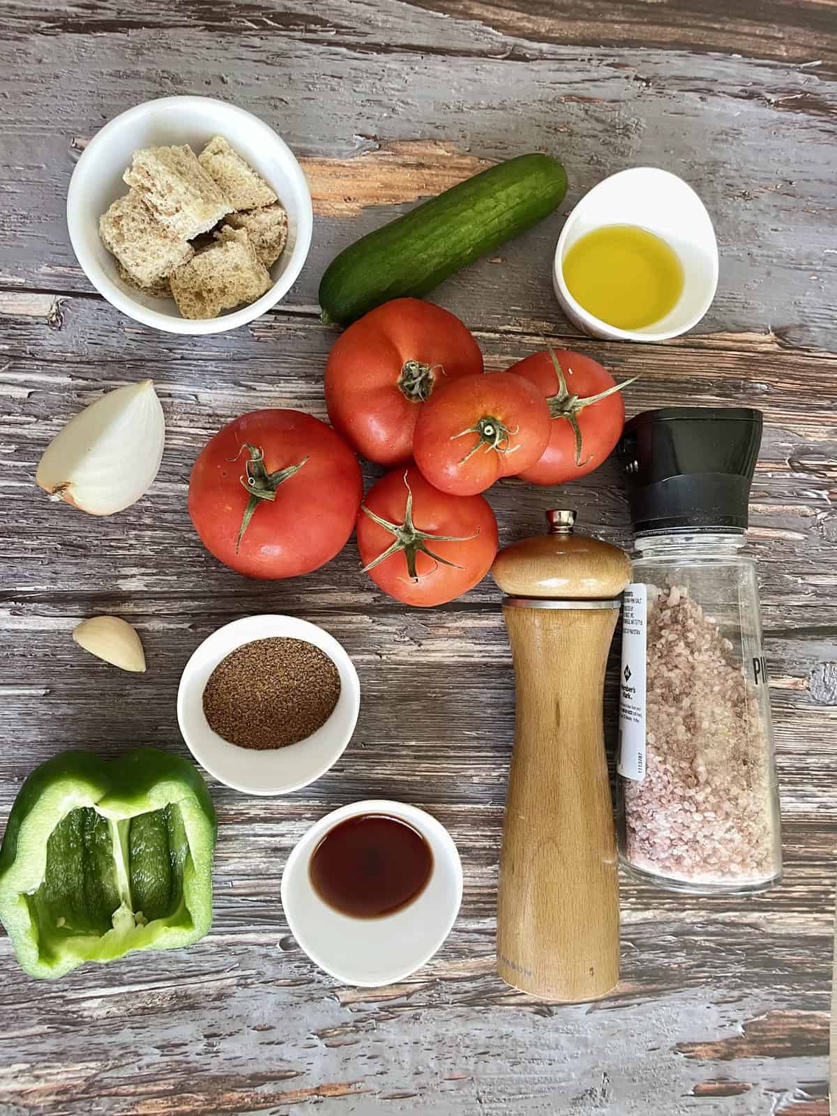 Ingredients for a traditional tomato and cucumber gazpacho laid out on a weathered wood table..