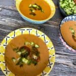 Gazpacho in three ceramic bowls on a weathered wood table garnished with chopped tomatoes, cucumber, chives and drizzled with olive oil.