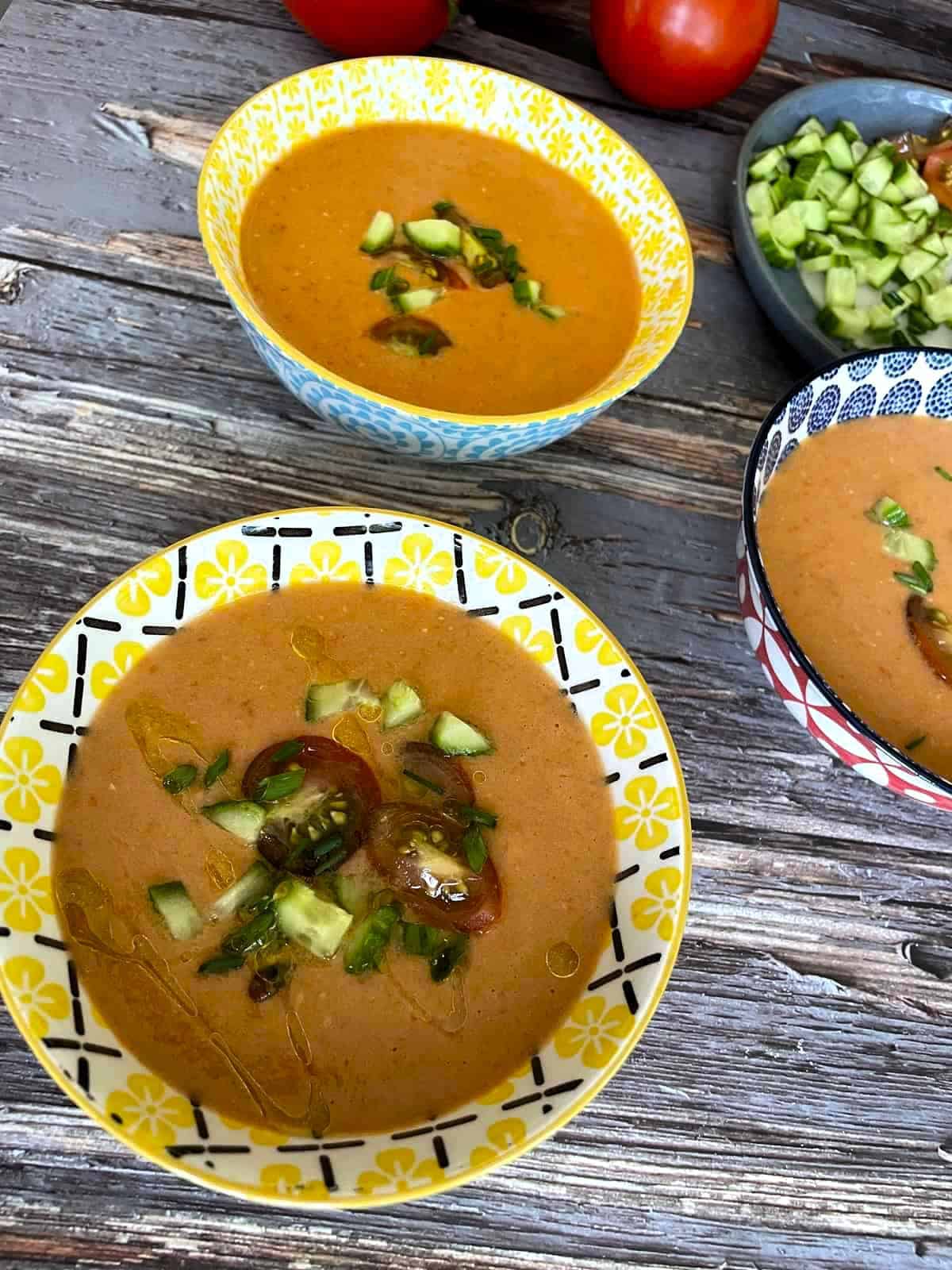 Gazpacho in three ceramic bowls on a weathered wood table garnished with chopped tomatoes, cucumber, chives and drizzled with olive oil.